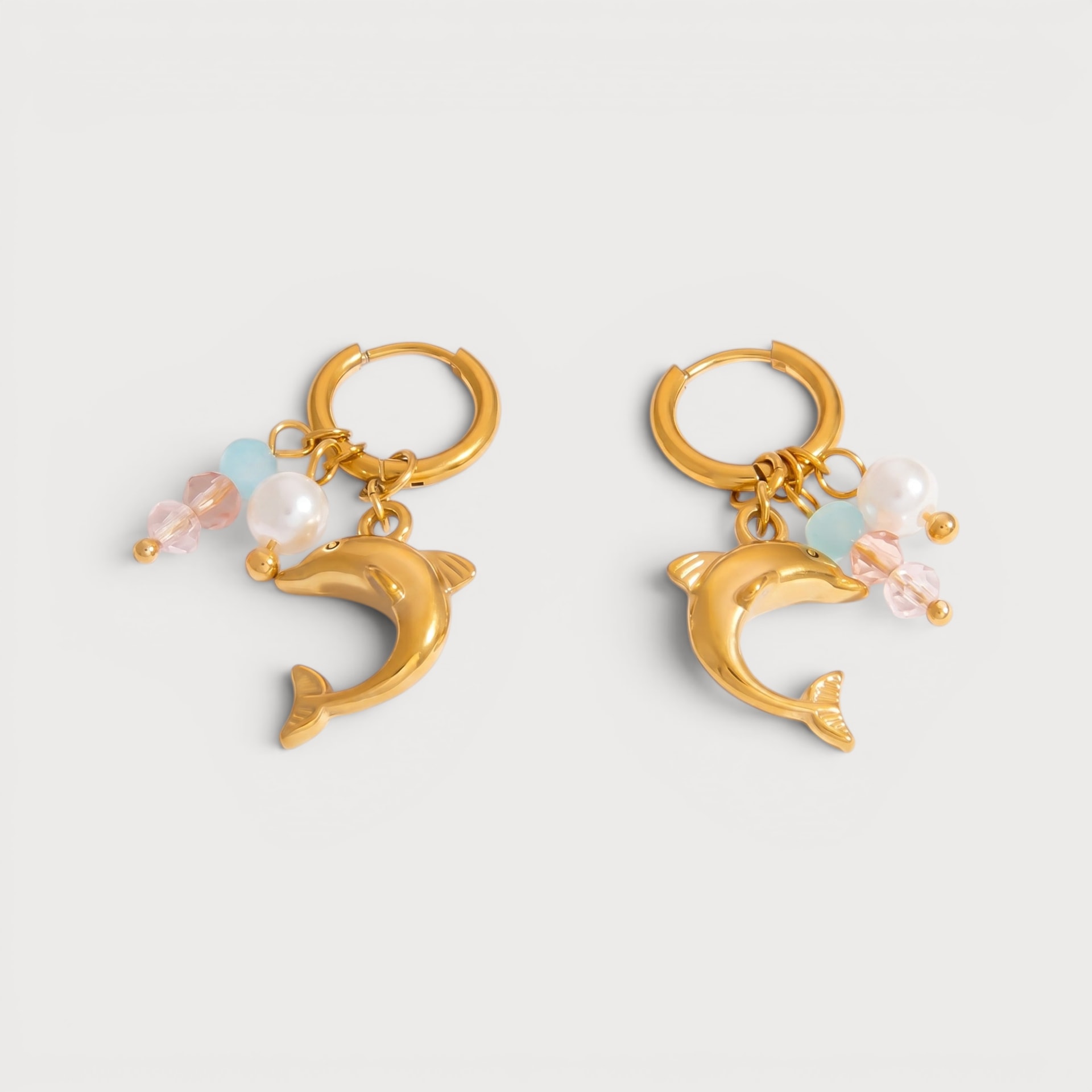 Boucles d’oreilles Manelle : des dauphins dorés sont suspendus aux créoles parmi des perles blanches et de petits éléments pastel.