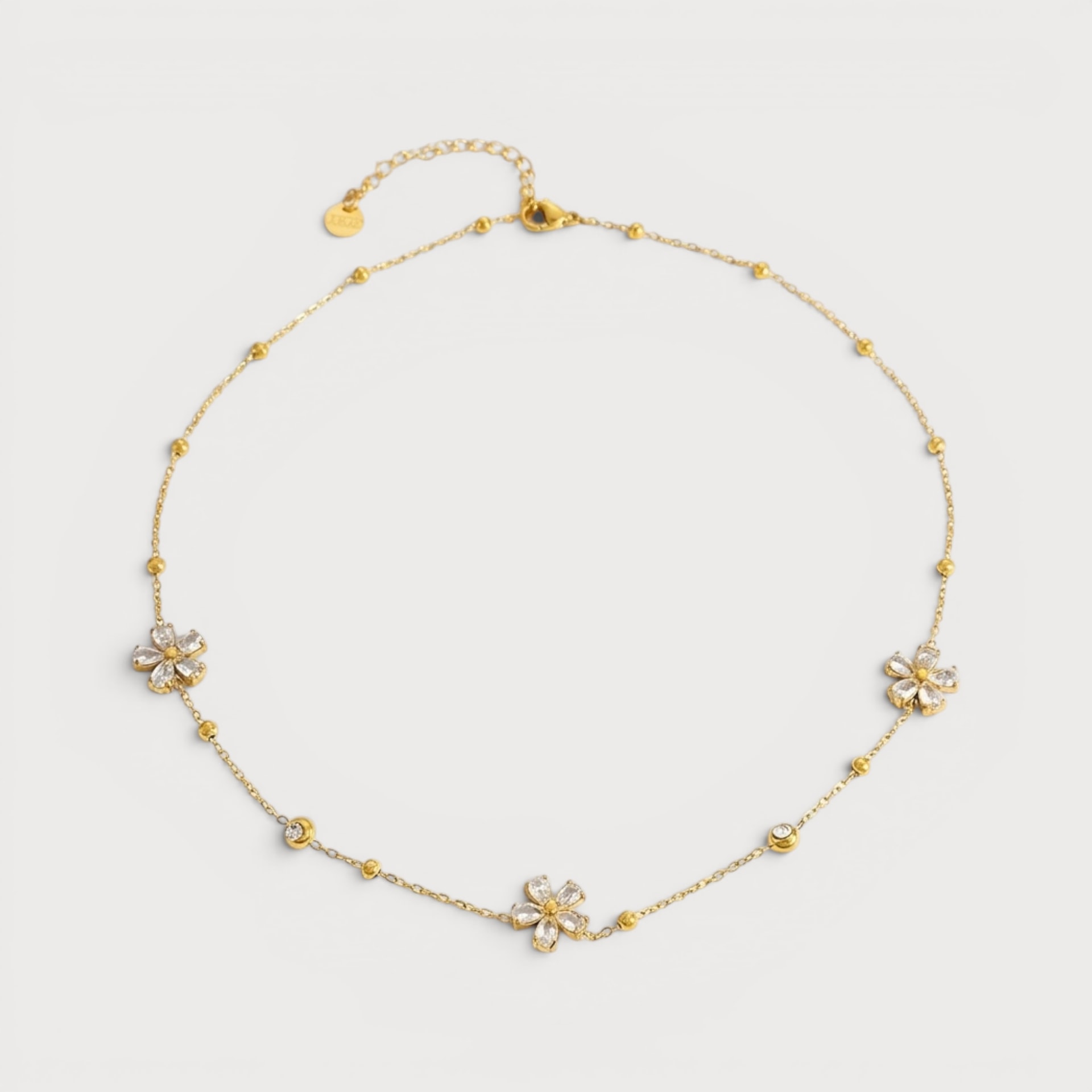 Collier Ghislaine : trois petits motifs floraux à pierres claires sont espacés sur une chaîne dorée ponctuée de petites boules.